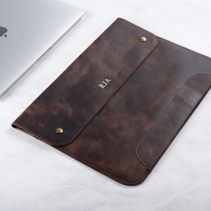 May include: A brown leather laptop sleeve with a flap closure and a brass button. The sleeve has a personalized monogram "RJA" in gold lettering.