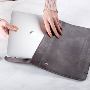 May include: A gray leather laptop sleeve with the name "Jonathan" embossed on the front. The sleeve is open and a silver laptop is partially inside.