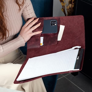 May include: A burgundy leather portfolio with multiple pockets, holding a smartphone, a notepad, and a pen. The portfolio is open, revealing the interior. The notepad has white lined paper. The portfolio is held by a person.