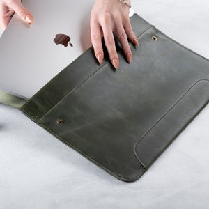 May include: A green leather laptop sleeve with a flap closure and a snap button. The sleeve is open and a silver laptop is partially inside.