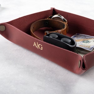 May include: A burgundy leather catchall tray with a watch, wireless earbuds, and cash inside. The tray has the initials "A/G" embossed on the front.