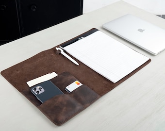 May include: A brown leather desk organizer with a notepad, pen, smartphone, and credit card. A silver laptop is also on the desk. The organizer has pockets for storage.