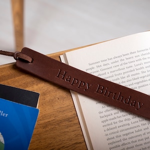 May include: A brown leather bookmark with the text "Happy Birthday" embossed on it. The bookmark is lying on top of an open book.
