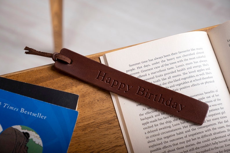May include: A brown leather bookmark with the text "Happy Birthday" embossed on it. The bookmark is lying on top of an open book.