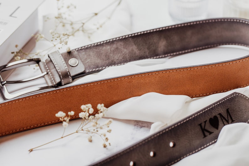 May include: Two leather belts, one brown and one gray, with a silver buckle. The gray belt has a heart and the initials "K & M" engraved on it.