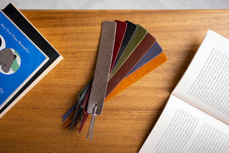 May include: A set of eight leather bookmarks in various colors, including gray, burgundy, olive green, brown, orange, and blue. The bookmarks are arranged in a fan shape on a wooden surface.