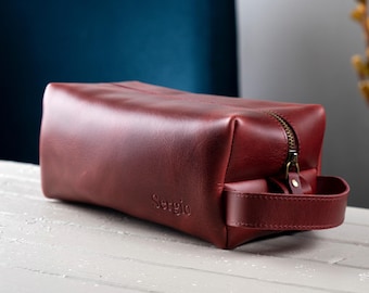 May include: A burgundy leather toiletry bag with a rectangular shape and a top zipper closure. The bag has a handle on the side and the name "Sergio" is embossed on the front. The bag is sitting on a white surface.