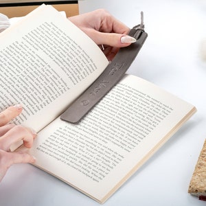 May include: A gray leather bookmark with the text "Ten More" engraved on it. The bookmark is being used to mark a place in a book.