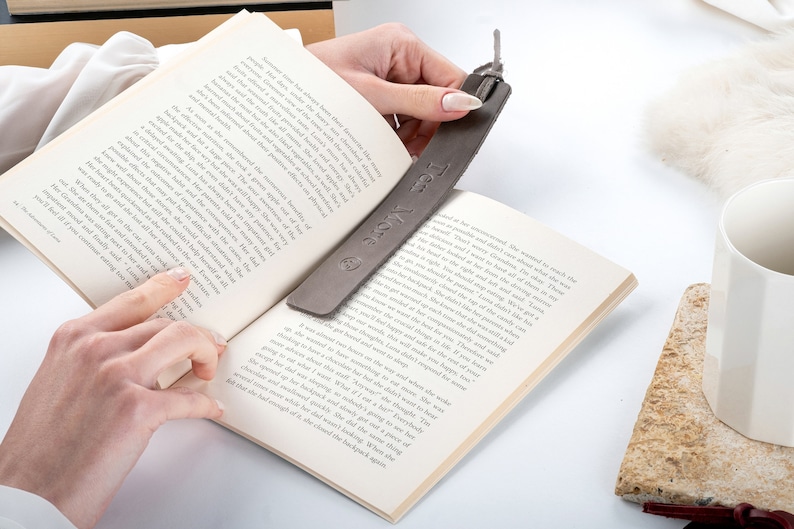 May include: A gray leather bookmark with the text "Ten More" engraved on it. The bookmark is being used to mark a place in a book.