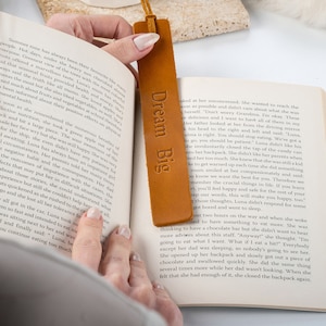 May include: A close-up of a brown leather bookmark with the text "Dream Big" embossed on it. The bookmark is being used to mark a page in a book.
