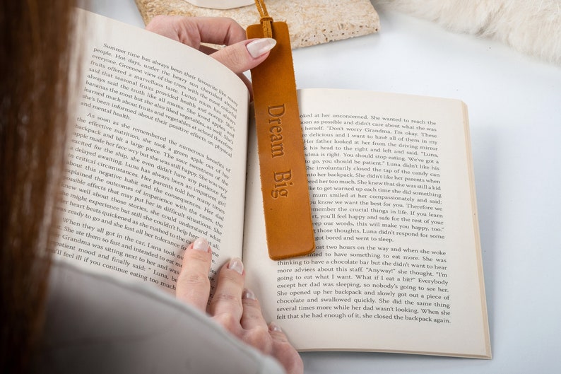 May include: A close-up of a brown leather bookmark with the text "Dream Big" embossed on it. The bookmark is being used to mark a page in a book.