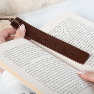 May include: A brown leather bookmark with the text "Happy Birthday" embossed on it. The bookmark is being used to mark a page in a book.