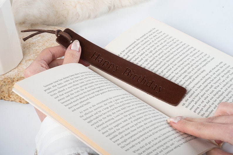 May include: A brown leather bookmark with the text "Happy Birthday" embossed on it. The bookmark is being used to mark a page in a book.