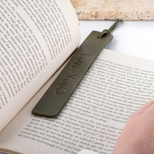 May include: A green leather bookmark with the text "Love You" embossed on it. The bookmark is lying on an open book.