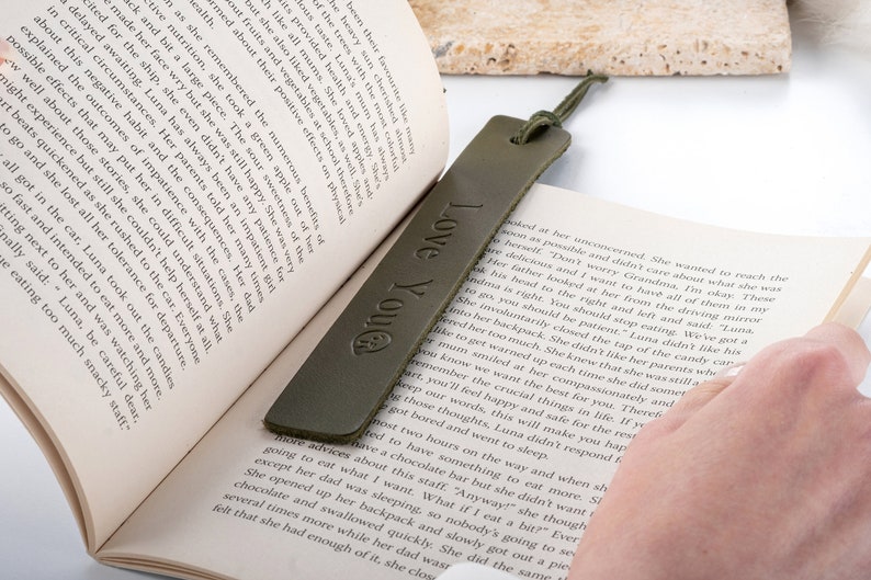 May include: A green leather bookmark with the text "Love You" embossed on it. The bookmark is lying on an open book.