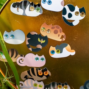 May include: A gold-coloured tray with a variety of colourful cat stickers. The stickers feature different cat breeds and poses, with each cat having sparkling gem eyes.