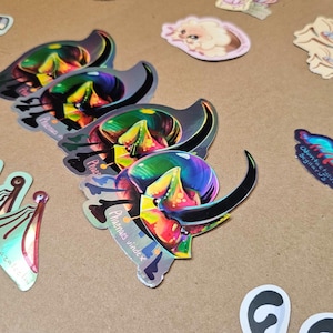 Puede incluir: Un conjunto de pegatinas iridiscentes de colores con escarabajos, mariposas y otros insectos. Las pegatinas son holográficas y tienen un acabado brillante. El texto en las pegatinas dice "Caught in Calisphlim" y "Adventures of Sigilar's Oddities".