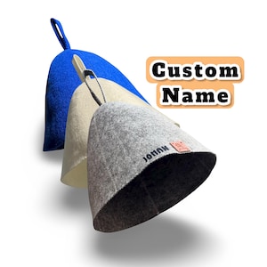 Könnte beinhalten: Drei Saunahüte aus Filz in Blau, Creme und Grau, jeweils mit einer Schlaufe zum Aufhängen. Der graue Hut trägt den Aufdruck "JOHAN" und ein kleines Lederetikett. Der Text "Custom Name" ist ebenfalls enthalten.