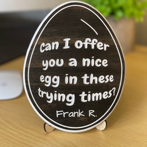 以下が含まれることがあります： 白い縁取りと白い文字で「Can I offer you a nice egg in these trying times?」と書かれた、卵型の木製看板。文字は遊び心のある丸みのあるフォントで書かれている。看板は木製スタンドの上に置かれている。看板の下部には「Frank R.」という文字が書かれている。