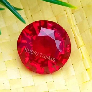 May include: A round, faceted, vibrant red gemstone with the text KUDRATGEMS printed in white across the center. The stone features a brilliant cut with multiple triangular and kite-shaped facets that reflect light, creating a deep, saturated crimson appearance.