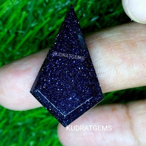 May include: A kite-shaped blue sandstone gemstone with a faceted surface and sparkling inclusions. The stone features the text KUDRATGEMS printed on its surface and is displayed against a bright green textured background.