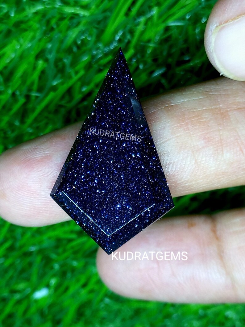May include: A kite-shaped blue sandstone gemstone with a faceted surface and sparkling inclusions. The stone features the text KUDRATGEMS printed on its surface and is displayed against a bright green textured background.