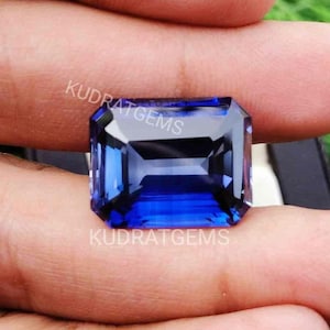 Può includere: Una pietra preziosa rettangolare a taglio smeraldo di un colore blu profondo e vibrante, con superfici sfaccettate che riflettono la luce. Il testo KUDRATGEMS è stampato in lettere bianche sulla superficie della pietra.
