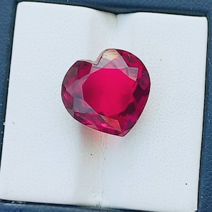 May include: A heart-shaped, deep red gemstone with a faceted cut. The gemstone is displayed on a white background.