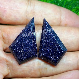 May include: Two shield-shaped blue sandstone gemstones with a faceted surface and sparkling metallic inclusions. The stones feature a deep navy blue color with a glittery appearance and the text KUDRATGEMS printed across them.