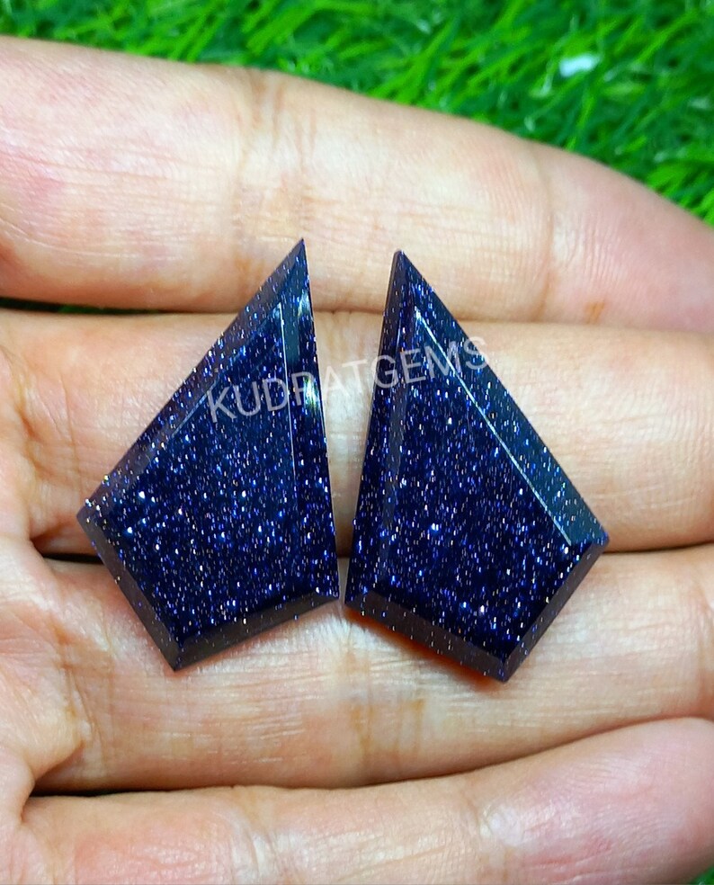May include: Two shield-shaped blue sandstone gemstones with a faceted surface and sparkling metallic inclusions. The stones feature a deep navy blue color with a glittery appearance and the text KUDRATGEMS printed across them.