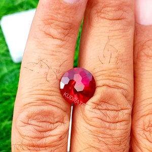 May include: A round, faceted, deep red gemstone with a brilliant cut. The surface of the stone features a geometric pattern of triangular and hexagonal facets that reflect light. The text KUDRATGEMS is printed in white on the lower portion of the stone.