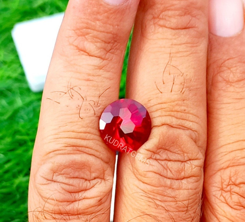 May include: A round, faceted, deep red gemstone with a brilliant cut. The surface of the stone features a geometric pattern of triangular and hexagonal facets that reflect light. The text KUDRATGEMS is printed in white on the lower portion of the stone.
