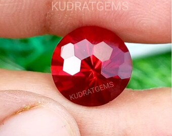 May include: A round, faceted, deep red gemstone with a brilliant cut and high clarity. The surface of the stone features the text KUDRATGEMS printed twice in white. The gem displays sharp geometric facets that reflect light across its surface.