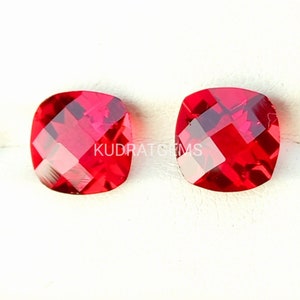 Op de afbeelding: Twee helderrode kussen geslepen edelstenen, elk met een gefacetteerd oppervlak, worden getoond op een wit oppervlak. De edelstenen zijn ongeveer 1 cm groot.