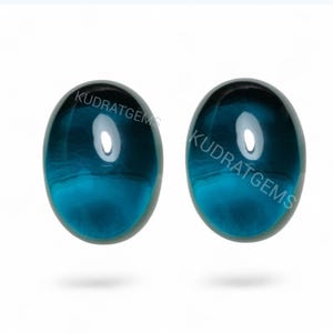 Op de afbeelding: Twee ovale, teal-kleurige edelstenen met een gepolijste, glanzende afwerking. De edelstenen hebben een doorschijnende kwaliteit, met een lichtere, bijna witte, ovale reflectie in het midden. De tekst "KUDRATGEMS" is op elke edelsteen gedrukt.