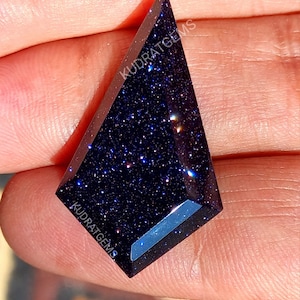 May include: A kite-shaped blue goldstone gemstone with a faceted surface and sparkling metallic inclusions. The dark blue stone features the text KUDRATGEMS printed on its surface in white lettering, showcasing a polished finish.