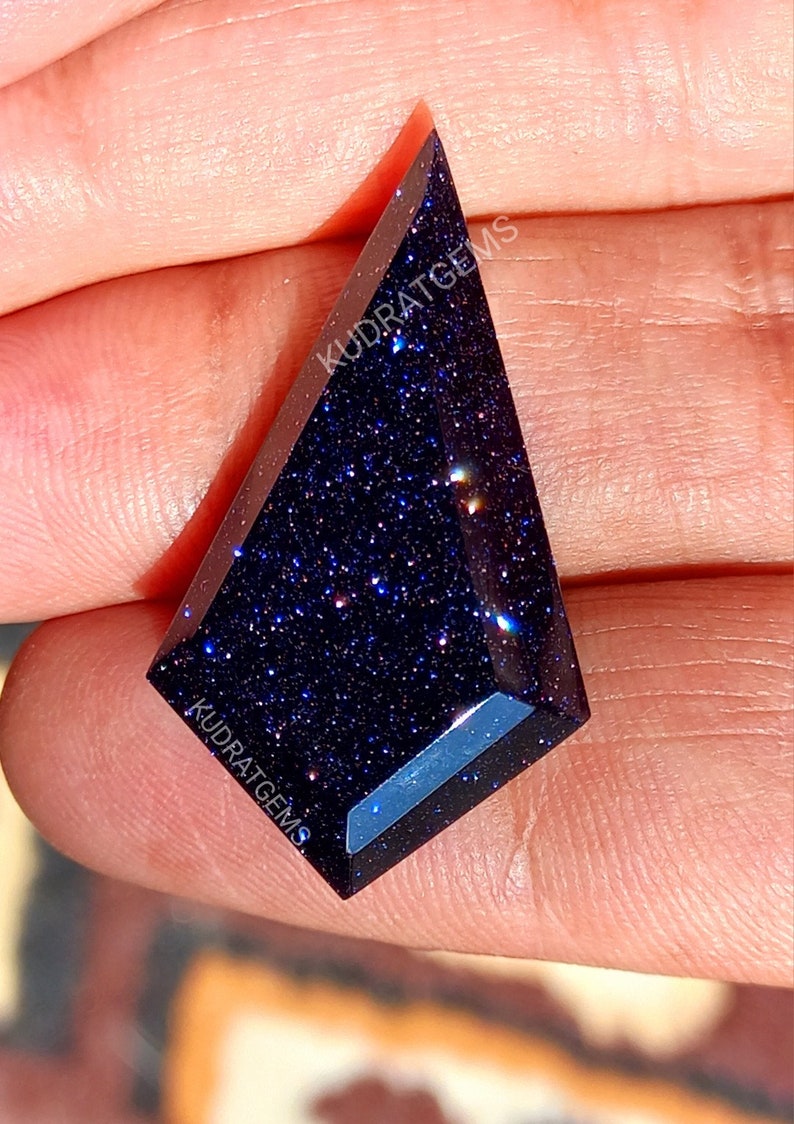 May include: A kite-shaped blue goldstone gemstone with a faceted surface and sparkling metallic inclusions. The dark blue stone features the text KUDRATGEMS printed on its surface in white lettering, showcasing a polished finish.