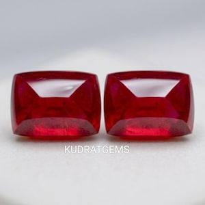 May include: Two cushion-cut, deep red gemstones with a high degree of clarity. The gems are displayed on a white surface. The text "KUDRATGEMS" is visible at the bottom of the image. These gems are suitable for jewelry making.