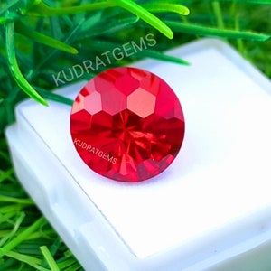 May include: A round, faceted, vibrant red gemstone with a brilliant cut, displaying geometric light reflections. The stone is placed on a white square display base. The text KUDRATGEMS is printed on the stone and above it in white.