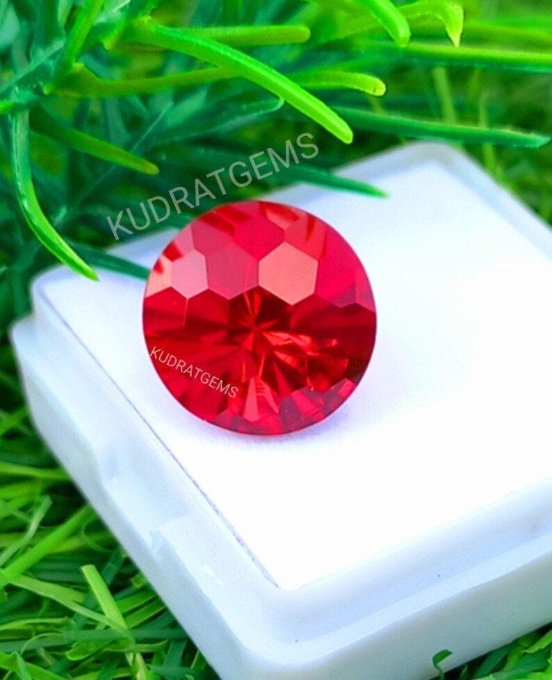 May include: A round, faceted, vibrant red gemstone with a brilliant cut, displaying geometric light reflections. The stone is placed on a white square display base. The text KUDRATGEMS is printed on the stone and above it in white.