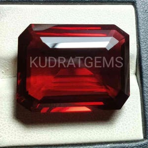 Könnte beinhalten: Ein tiefroter Edelstein mit einem rechteckigen Smaragdschliff. Der Edelstein ist in einer weißen Ringfassung gefasst. Der Text "KUDRATGEMS" ist im Bild sichtbar.