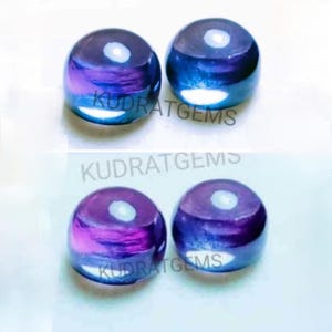 Op de afbeelding: Vier ronde cabochon edelstenen met een glad, gepolijst oppervlak. De stenen vertonen een verloop van dieppaarse en blauwe tinten met een reflecterende lichtvlek op elk exemplaar. De tekst KUDRATGEMS is zichtbaar op de stenen.