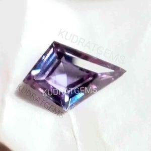 Könnte beinhalten: Ein drachenförmiger facettierter Edelstein mit einem Farbwechsel von Violett zu Blau. Der Stein hat eine quadratische Facette in der Mitte, die von dreieckigen Facetten umgeben ist. Der Text KUDRATGEMS ist auf dem Edelstein sichtbar.