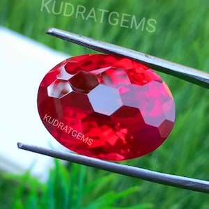 Puede incluir: Una gema ovalada de color rojo vibrante con una superficie facetada sostenida por pinzas de metal. La gema presenta un patrón de corte hexagonal distintivo en la parte superior. El texto KUDRATGEMS es visible dos veces en letras blancas.