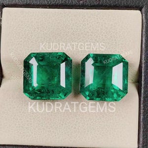 May include: Two vibrant green emerald-cut gemstones displayed in a jewelry box. The gems are a rich, deep green color with a rectangular shape. The text "KUDRAT GEMS" is printed on the box.