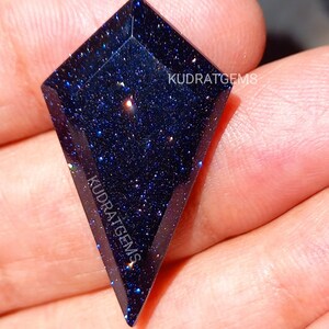 May include: A kite-shaped blue goldstone gemstone with a faceted surface and a deep navy blue color filled with sparkling metallic flecks. The text KUDRATGEMS is printed on the surface of the stone in a white sans-serif font.