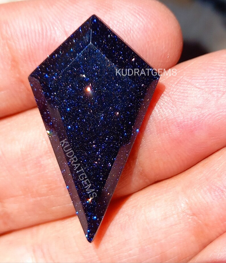 May include: A kite-shaped blue goldstone gemstone with a faceted surface and a deep navy blue color filled with sparkling metallic flecks. The text KUDRATGEMS is printed on the surface of the stone in a white sans-serif font.