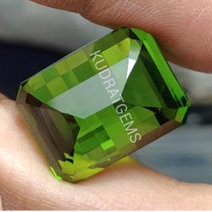 May include: A faceted, rectangular gemstone in shades of green is held between fingers. The gem has the text "KUDRAT GEMS" etched on its surface. The gem's cut creates multiple reflective surfaces.