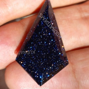 May include: A kite-shaped blue goldstone gemstone with a faceted surface and sparkling metallic inclusions. The dark blue stone features a geometric cut and is marked with the text KUDRATGEMS repeated across the surface.