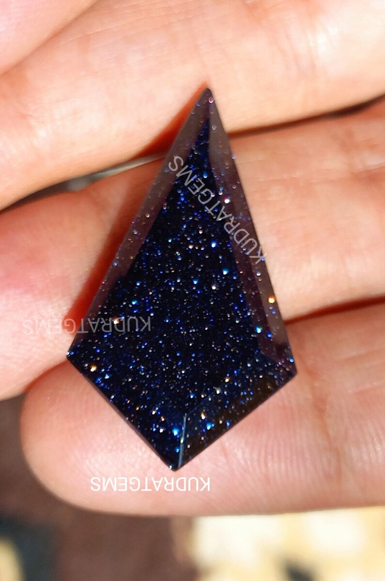 May include: A kite-shaped blue goldstone gemstone with a faceted surface and sparkling metallic inclusions. The dark blue stone features a geometric cut and is marked with the text KUDRATGEMS repeated across the surface.